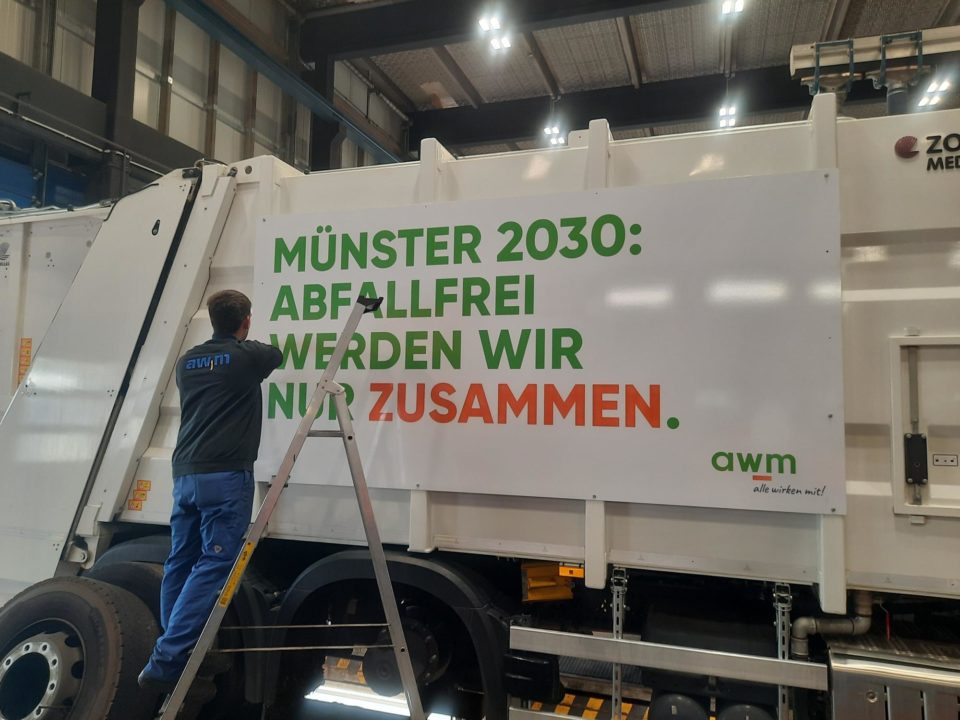 Mitarbeiter der awm schraubt eine Platte mit der Aufschrift "Münster 2030: Abfallfrei werden wir nur zusammen" an einem Abfallsammelfahrzeug an.